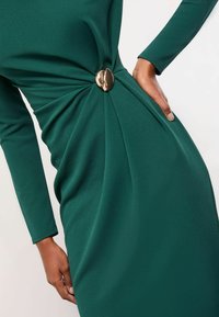 Žalios spalvos, ilgomis rankovėmis suknelė, pagaminta iš glotnaus audinio, su suktos juosmens detale ir blizgiu auksiniu dekoratyviniu mygtuku. Paprastas ir elegantiškas dizainas.