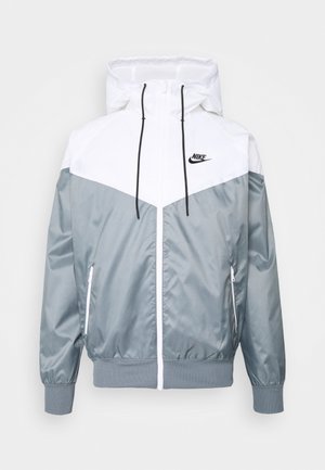 Biała i szara kurtka przeciwdeszczowa Nike z kapturem, czarnymi sznurkami, zamkiem błyskawicznym z przodu, zamykanymi kieszeniami bocznymi i logo Nike na piersi.