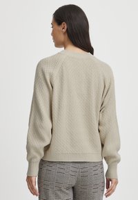 Pull en beige texturé avec des manches raglan et un col côtelé. Porté sur un pantalon à carreaux, montrant une coupe ample et un motif en maille.