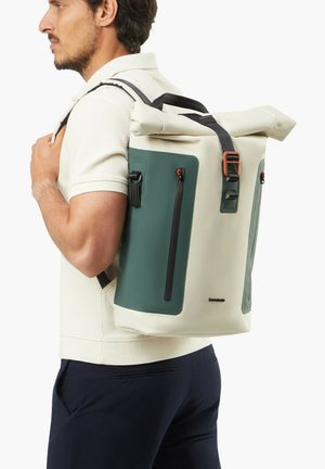 Mann trägt einen cremefarbenen und grünen Rolltop-Rucksack mit vorderen vertikalen Reißverschlusstaschen und schwarzen Trägern über einem cremefarbenen Hemd.