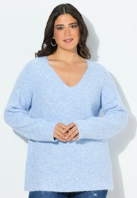 Frau mit dunklem, welligem Haar, die einen hellblauen, flauschigen V-Ausschnitt-Pullover und Ohrreifen trägt, mit leicht gefalteten Händen, vor einem weißen Hintergrund stehend.