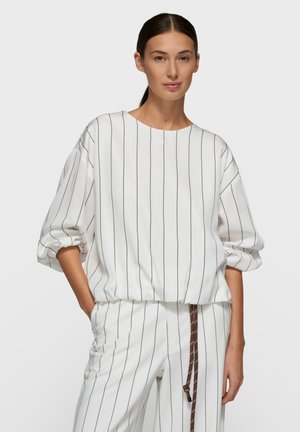 Kobieta ubrana w biały strój z cienkimi pionowymi czarnymi paskami, luźna bluzka z długim rękawem i spodnie z brązowym sznurowaniem, pozująca z jedną ręką w kieszeni.