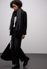 Manteau noir sur un pull léger, associé �à un jean évasé foncé et des bottines noires. Une écharpe noire texturée avec des franges apporte une touche de détail.