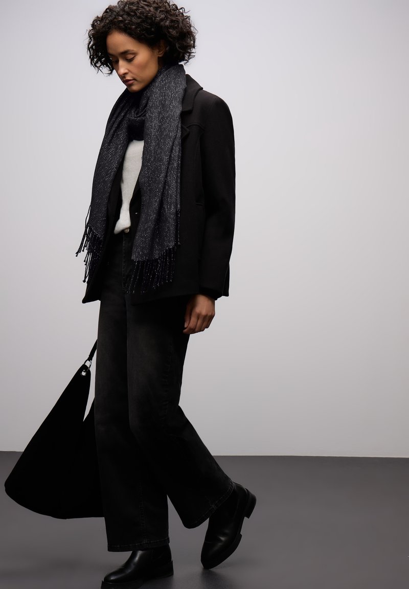 Manteau noir sur un pull léger, associé à un jean évasé foncé et des bottines noires. Une écharpe noire texturée avec des franges apporte une touche de détail.