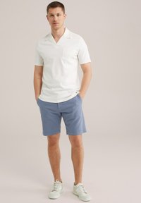 Polo alb cu maneci scurte și buzunar pe piept, asortat cu pantaloni scurți bleu deschis și adidași albi. Textură netedă, design casual.