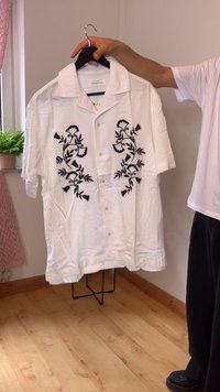 Camisa branca de manga curta com bordado floral preto, com botões na frente e colarinho aberto. Tecido texturizado com um corte descontraído.