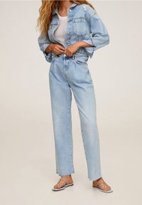 Žena v lehké modré džínové bundě a džínech s vysokým pasem a rovnými nohavicemi, bílém pleteném topu a sandálech na plochém podpatku se stahovacími pásky.