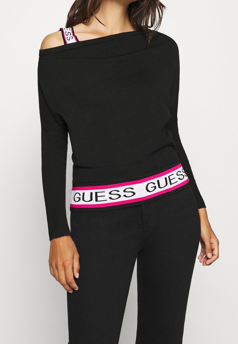 Maglione nero off-shoulder con maniche lunghe, caratterizzato da una vita in contrasto rosa e bianco con il logo "GUESS" e una texture a coste.