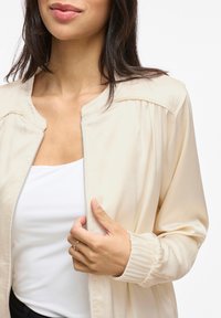 Ljust beige zip-jacka med en mjuk yta, plisserade axlar och elastiska manschetter. Bärs över en vit topp med rund halsringning.