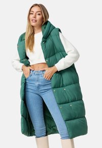 Gepolsterte grüne Weste mit Kapuze, ausgestattet mit Druckknöpfen und im langen Design. Trägt man über einem weißen, kurzen Pullover und blauen Jeans.