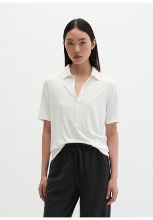 Jeune femme aux longs cheveux raides porte une chemise blanche à manches courtes boutonnée rentrée dans un pantalon noir à cordon, debout devant un fond uni.