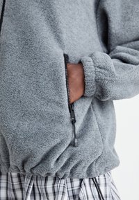 Graue Fleecejacke mit einer Reißverschlusstasche, elastischen Bündchen und einem Kordelzugsaum. Das strukturierte Material bietet Wärme und zeichnet sich durch ein lässiges Design aus.