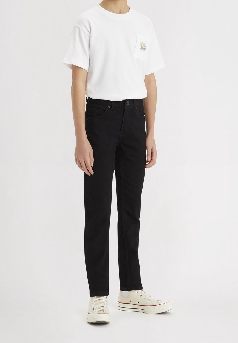 T-shirt en coton blanc avec détail pochette, associé à un jean en denim noir. Les pieds sont chaussés de baskets couleur crème, avec une finition en toile texturée.