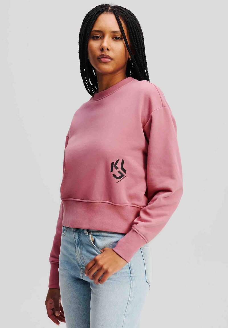 Felpa cropped rosa con scollatura rotonda e maniche lunghe. Presenta un logo nero sul lato sinistro e una texture morbida. Abbinata a jeans azzurri chiari.
