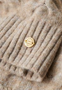 Suéter de lana beige con textura de canalé; presenta una etiqueta redonda dorada grabada con "Super Dry Premium NY." Apariencia suave y acogedora.