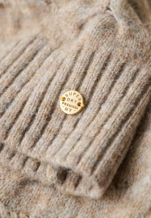 Pull en laine beige avec texture côtelée ; présente une étiquette ronde en or embossée portant l'inscription "Super Dry Premium NY." Apparence douce et confortable.