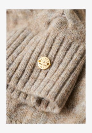 Pull en laine beige avec texture côtelée ; présente une étiquette ronde en or embossée portant l'inscription "Super Dry Premium NY." Apparence douce et confortable.