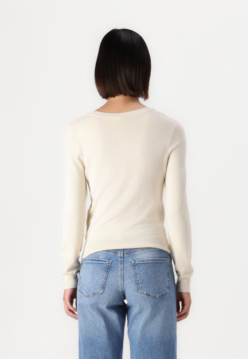 Maglione color crema, maniche lunghe, realizzato in materiale morbido, con scollatura rotonda e polsini a coste, abbinato a jeans azzurri chiari.