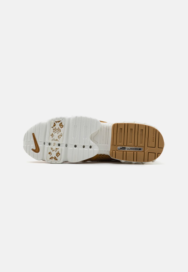 La semelle extérieure de la sneaker Nike présente une semelle intermédiaire blanche avec un grip en caoutchouc à motifs, des accents dorés et un dessous texturé de couleur beige.