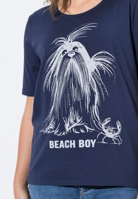 Námořnická modrá tričko z měkké tkaniny s bílou grafikou chlupaté bytosti a kraba, pod níž je napsáno "BEACH BOY".