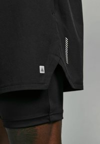 Shorts de sport noirs avec une fente sur le côté, étiquette de marque et détails en rayures subtiles sur le côté. Tissu texturé avec une finition lisse.