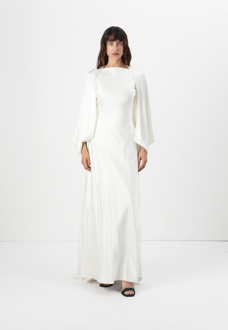 Robe blanche longue jusqu'au sol avec un corsage ajusté et de larges manches tombantes. Texture lisse, détails minimaux, portée avec des chaussures noires à bout ouvert.
