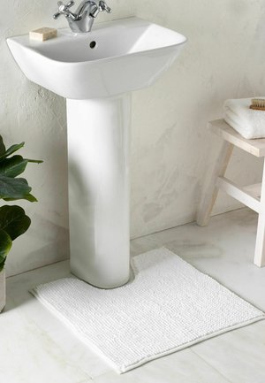 Weißes Standwaschbecken mit Chromarmatur, beiger Seifenstück, weiße U-förmige Badematte, grüne Pflanze und hocker aus Holz mit gefalteten Handtüchern.