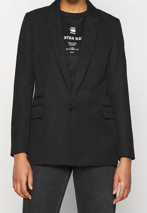 Person iført en tætsiddende sort blazer over en sort t-shirt med grafik og mørkegrå jeans, stående mod en ensartet lys baggrund.