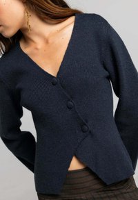 Cárdigan de punto azul marino con escote en V profundo, tres botones y dobladillo asimétrico. Textura suave y mangas largas con detalles acanalados.