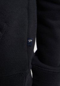 Marineblauwe sweatshirt met een zachte textuur, geribde manchetten en een klein logopatch met witte borduring op een donkerblauwe achtergrond.