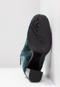 Bottine en velours vert avec un talon texturé noir. Détail de zip latéral et logo "TOMMY JEANS" embossé sur le talon pour le branding.