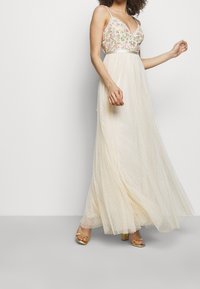 Maxi šaty s květinově vyšívaným živůtkem, výstřihem do V a průhlednou sukní s puntíky v krémové barvě. Mají tenké ramínka a přiléhavý pas. Viditelné zlaté podpatky.