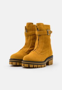 Bottines en daim jaune avec une semelle plateforme noire épaisse, dotées de zips latéraux et d'une sangle avec une boucle en métal.
