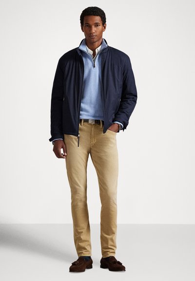 Vīrietis, tērpies tumši zilā žaketē pār gaiši zilu kvadrātveida rāvējslēdzēja džemperi, bēšās biksēs un brūnās kurpēs ar bārkstīm, stāv pret vienveidīgu baltu fonu.