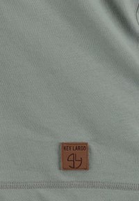 Tissu en coton gris avec une patch en cuir brun portant l'inscription "KEY LARGO" en relief. Texture lisse avec un design simple et sans embellissement.