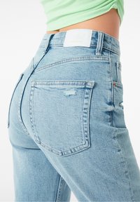 Ljusblå denimjeans med hög midja, som har två bakfickor och en diskret slitning. Tyget har en slät, lätt texturerad yta.