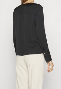 Blusa nera in satin con maniche lunghe, caratterizzata da un collo rotondo e dettagli con bottoni sui polsini, abbinata a pantaloni chiari.
