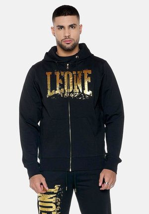 Uomo che indossa una felpa nera con cappuccio e pantaloni abbinati con scritta dorata usurata "LEONE 1947", in piedi contro uno sfondo chiaro e uniforme.