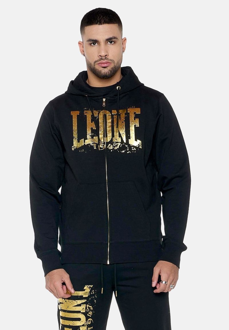 Homme portant un sweat à capuche noir et un pantalon assorti avec un texte doré usé « LEONE 1947 », debout devant un fond clair uni.