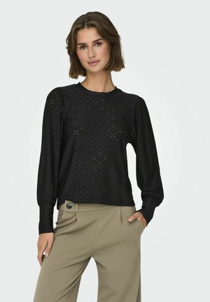 Svart långärmad topp med ett texturerat ögla-mönster, rund halsringning och puffärmar, kombinerad med ljus khaki-byxor.