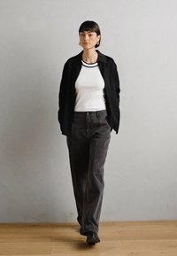 Zwarte button-up shirt over een witte ribbed tanktop, gecombineerd met hooglopende grijze corduroy broek en zwarte schoenen, op een houten vloer.