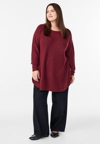 Burgundijas krāsas pārmēru džemperis ar garām piedurknēm, sānu pogu akcenti, apvienots ar melnām platām biksēm un melniem rotātiem apaviem.