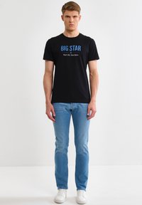 Giovane uomo che indossa una T-shirt nera con il logo "BIG STAR", jeans azzurri e sneakers bianche, in piedi contro uno sfondo bianco uniforme.