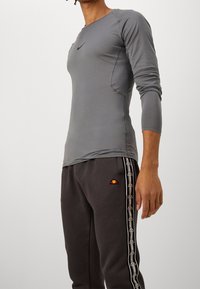 Man wearing fitted long-sleeve gray Nike shirt and dark gray Ellesse sweatpants with branded side stripes, standing against plain background.