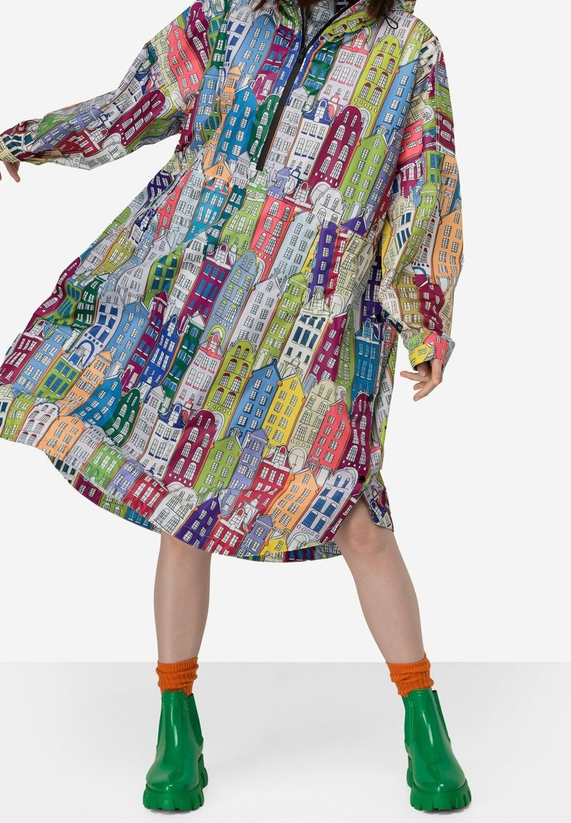 Meerkleurige jurk met een stadsgezichtsprint, oversized mouwen, een halve rits aan de voorkant en een losse pasvorm. Draagt groene laarzen en oranje sokken.