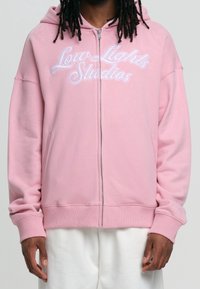 Sweat à capuche zippé rose avec capuche à cordon de serrage. Porte le texte "Low Lights Studios" en blanc, avec des poignets et un ourlet côtelés. Matière en coton doux.
