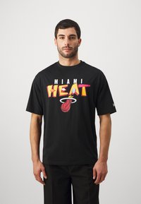 Camiseta de algodón negra con un gráfico llamativo de "MIAMI HEAT" en rojo, amarillo y blanco, junto con un logo de baloncesto. Manga corta y corte clásico.