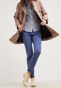 Trench-coat beige clair, chemise à rayures bleues, pantalon bleu ajusté et bottines crème. Tissu lisse et design soigné.