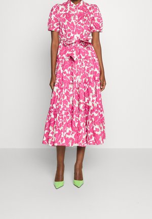 Kobieta ubrana w różowo-białą kwiatową sukienkę midi z bufkami na rękawach i wiązaniem w talii, w zestawieniu z zielonymi szpiczastymi szpilkami.