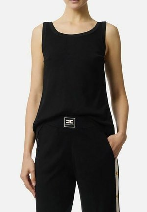 Donna con canotta nera senza maniche e pantaloni neri con striscia bianca e patch logo in vita, in piedi con la mano in tasca.